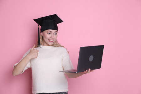Happy student with laptop showing thumbs up after graduation on pink backgroundの写真素材