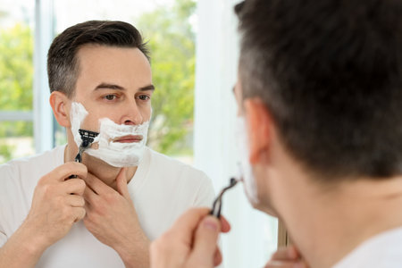 Handsome man shaving with razor near mirror at homeの写真素材