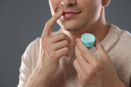 Young man applying lip balm onto his lips on grey background, closeupの写真素材