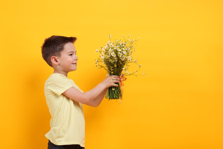 Cute little boy giving bouquet of beautiful chamomile flowers on yellow background. Space for textの写真素材