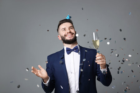 Happy Birthday. Handsome man with party hat and glass of champagne under confetti shower on grey backgroundの写真素材