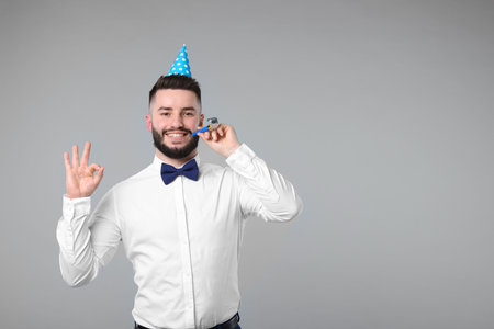 Happy Birthday. Handsome man with party hat and blower showing ok gesture on grey background, space for textの写真素材