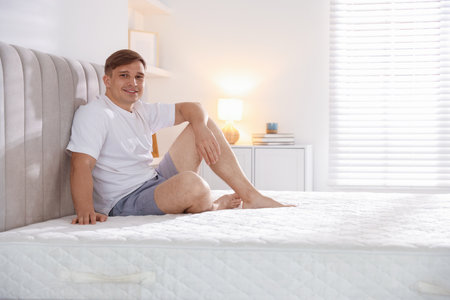 Young man sitting on bed with soft mattress at home, space for textの写真素材