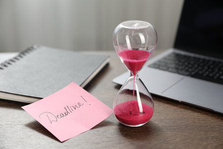 Sticky note with word Deadline, hourglass, notebook and laptop on wooden table indoors, closeupの写真素材