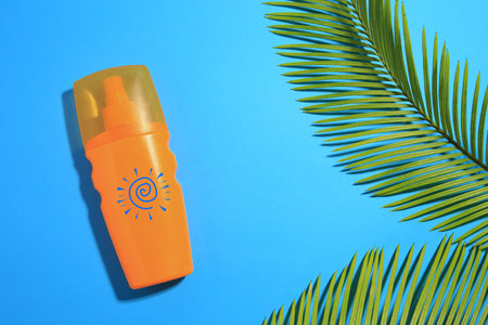 Sunscreen product and green palm leaves on blue background, top view. Bottle with drawing of sunの写真素材
