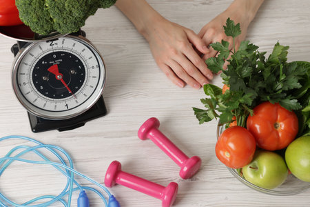 Woman at light wooden table with healthy food, kitchen scale, skipping rope and dumbbells, top viewの写真素材