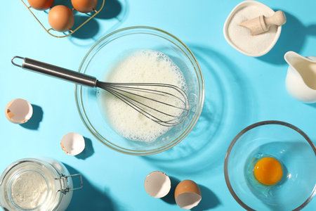 Whisk, egg whites in bowl and other products on light blue table, flat lay