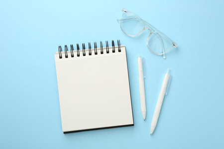 Notebook, glasses and pens a on light blue background, flat lay