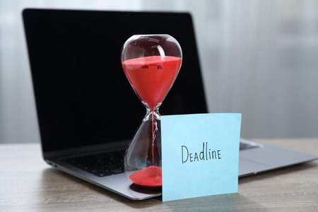 Sticky note with word Deadline, hourglass and laptop on wooden table indoors, closeup