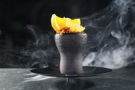 Hookah bowl with citrus fruits and smoke on black table, closeup