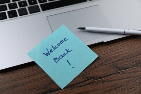 Paper note with words Welcome Back, laptop and pen on wooden table, closeupの写真素材