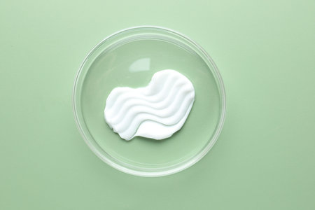 Cosmetic product in Petri dish on green background, top view