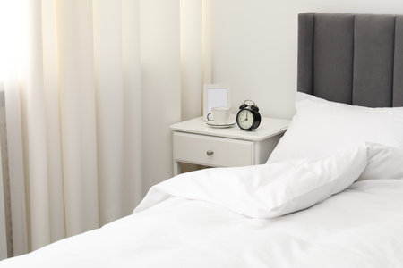 Bedside table with alarm clock, coffee and frame near bed indoors