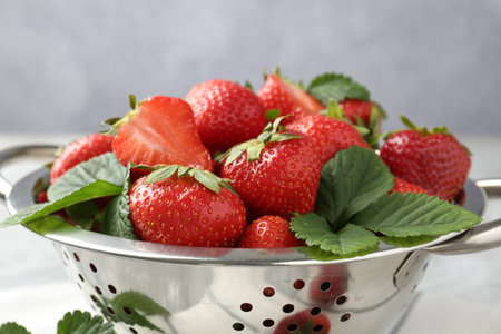 Delicious ripe strawberries and green leaves in colander on table, closeupの写真素材