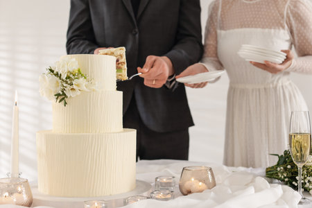 Groom taking piece of wedding cake while bride holding plates at table indoorsの写真素材