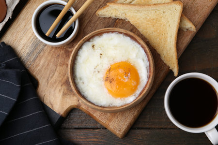 Half-boiled egg in bowl, soy sauce, chopsticks, coffee and toasted bread on wooden table, flat lay....