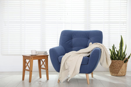 Comfortable blue armchair, blanket, side table and houseplant near white wall indoorsの写真素材