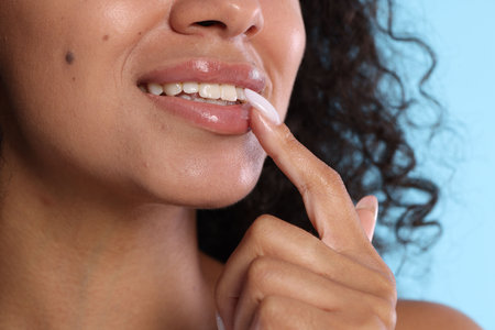 Smiling woman applying lip balm onto her lips on light blue background, closeup
