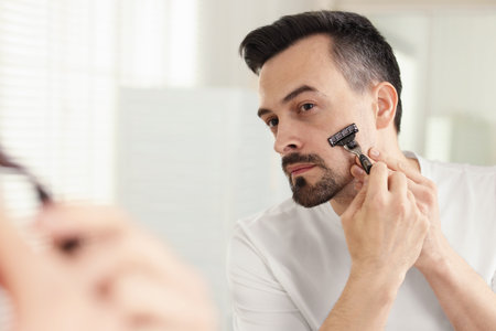 Handsome man shaving with razor near mirror at homeの写真素材