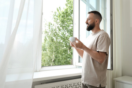 Harmony. Man with cup of drink near window at homeの写真素材