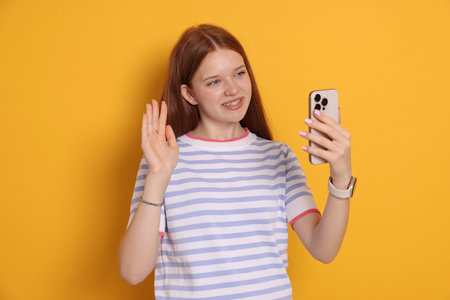 Smiling teenage girl having videocall by smartphone on yellow backgroundの写真素材