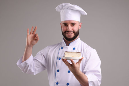 Happy confectioner in uniform holding piece of delicious cheesecake and showing ok gesture on light grey backgroundの写真素材