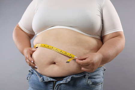 Overweight woman measuring her belly with tape on grey background, closeupの写真素材