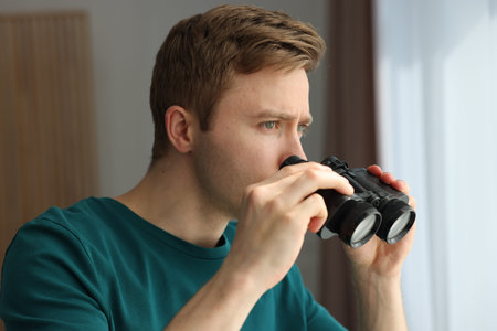 Paranoia. Worried man with binoculars near window indoors