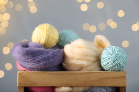 Soft balls of yarn and felting wool in crate against gray background with blurred lights, closeup