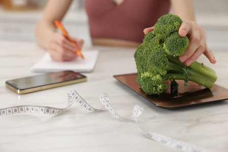 Woman developing diet plan while weighing broccoli on kitchen scale at white marble table indoors, closeupの写真素材