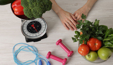 Woman at light wooden table with healthy food, kitchen scale, skipping rope and dumbbells, top viewの写真素材
