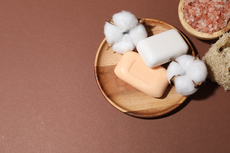 Soap bars, sea salt, cotton flowers and loofah sponge on brown background, flat lay. Space for textの写真素材