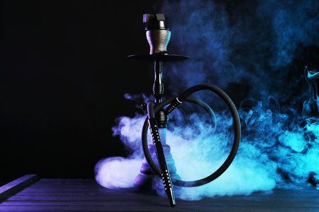Hookah on wooden table with smoke in color lights against black backgroundの写真素材