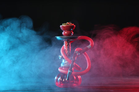 Hookah on wooden table with smoke in color lights against black background
