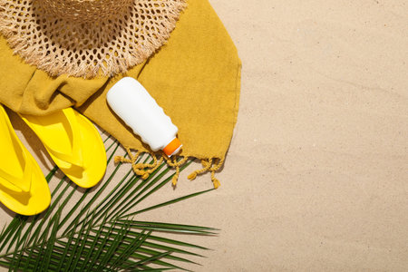 Beach towel, straw hat, flip flops, sunscreen product and palm leaf on sand outdoors, flat lay....