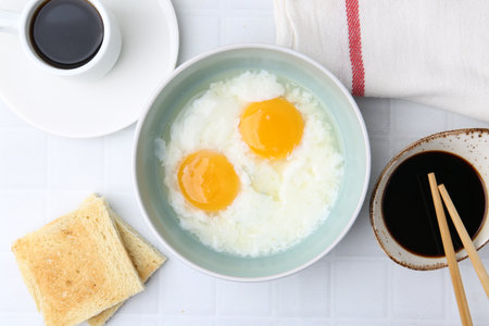 Traditional asian breakfast with half-boiled eggs in bowl, soy sauce, bread, chopsticks and coffee on white tiled table, flat layの写真素材