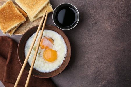 Traditional Asian breakfast with half-boiled eggs in bowl, chopsticks, soy sauce and toasted bread on brown table, flat lay. Space for textの写真素材