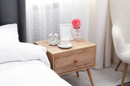 Bedside table with coffee, flower and alarm clock near bed indoors