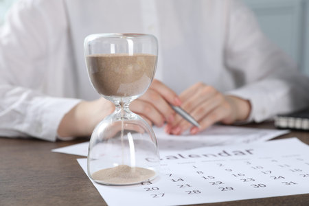 Deadline. Woman with calendar and pen at wooden table in office, focus on hourglass
