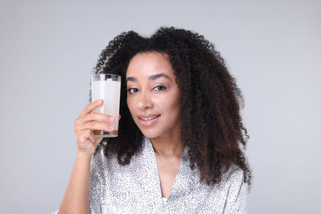 Beautiful Brazilian woman with glass of collagen water on gray background