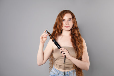 Beautiful woman with long wavy hair and curling iron on grey background, space for textの写真素材