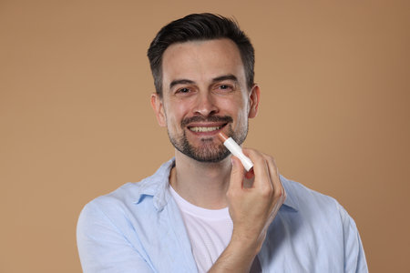 Smiling man applying lip balm onto his lips on light brown backgroundの写真素材