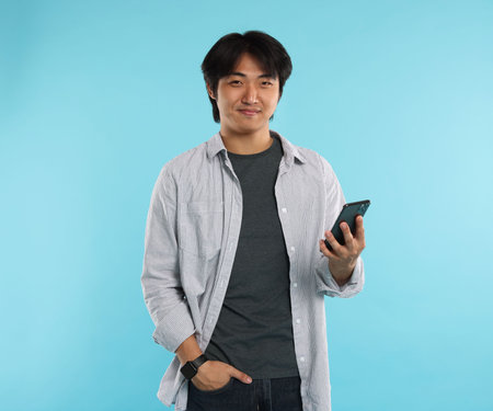 Handsome Asian man with smartphone on light blue background