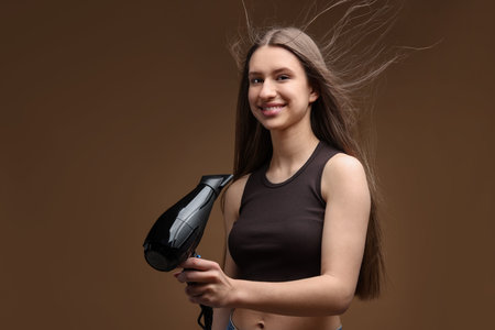 Smiling teenage girl with hair dryer on brown backgroundの写真素材