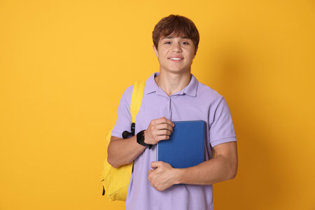 Smiling teenage boy with book and backpack on yellow backgroundの写真素材