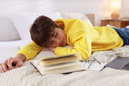 Tired teenage student sleeping near books and laptop on bed at homeの写真素材