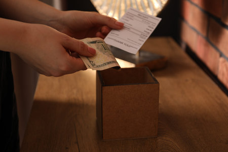 Woman with receipt putting tips into box at wooden table indoors, closeup