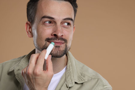 Handsome man applying lip balm onto his lips on light brown backgroundの写真素材