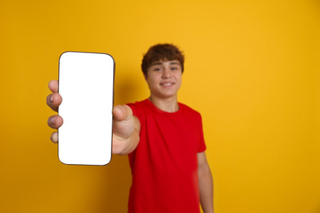 Smiling teenage boy showing smartphone with blank screen on yellow background, selective focus. Mockup for designの写真素材