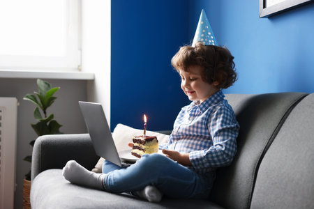 Birthday celebration. Little boy with piece of cake having video call via laptop on sofa at homeの写真素材